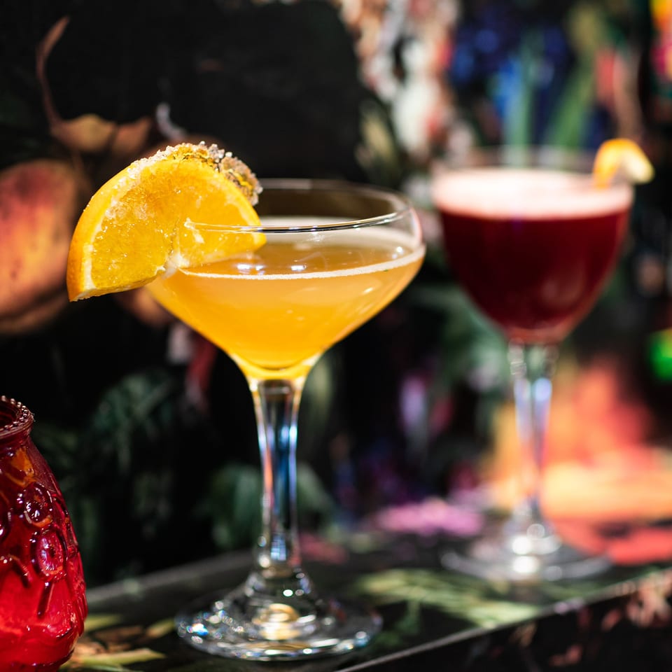 Orange cocktail in a sugar-rimmed coupe glass, surrounded by colorful drinks on a dimly lit bar counter.