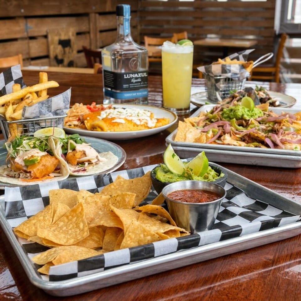 Mexican restaurant spread with tacos, loaded nachos, fries, enchiladas, guacamole, salsa, a citrus drink, and tequila on a wooden table.