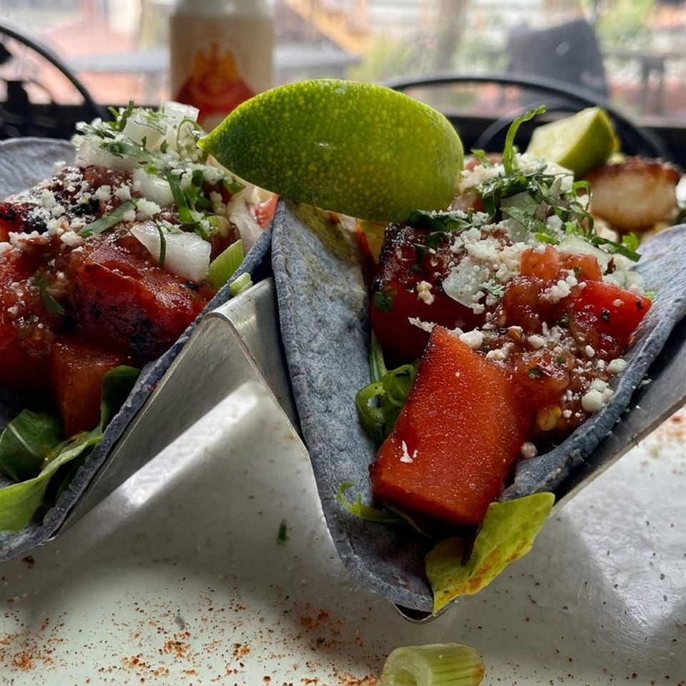 Two blue corn tacos filled with grilled watermelon, cotija cheese, cilantro, onions, and lime wedges on a metal stand.