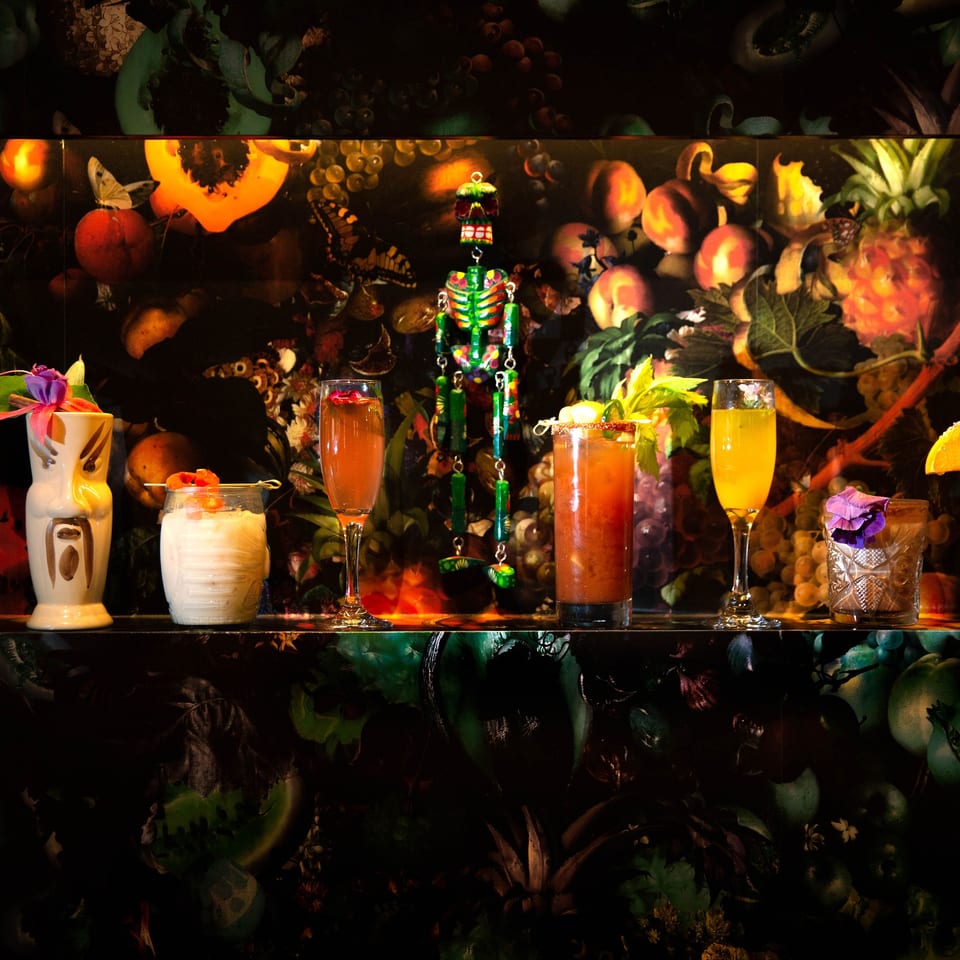 Colorful tropical cocktails arranged on a shelf with tiki glassware, fruit, flowers, and a decorative skeleton centerpiece.