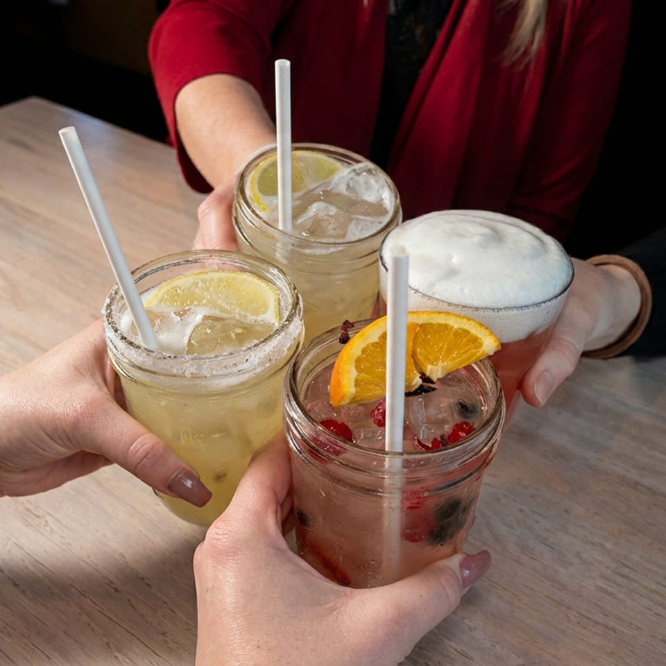 Four colorful cocktails in mason jars with citrus garnishes and straws are raised together over a wooden restaurant table.