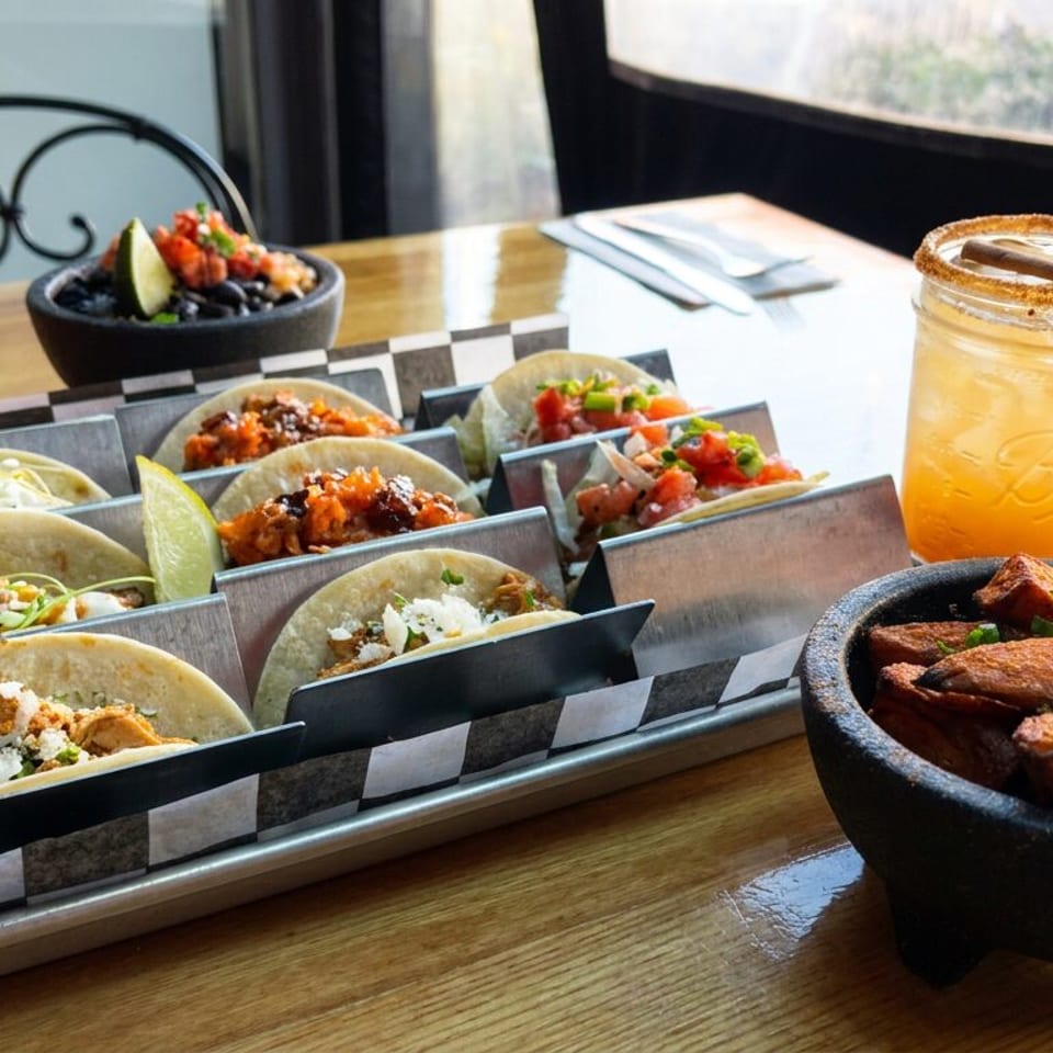 Tray of assorted tacos with lime wedges, a bowl of roasted vegetables, and two colorful drinks on a restaurant table.