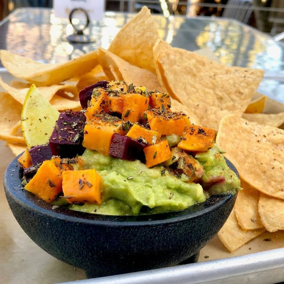Guacamole topped with roasted orange and purple vegetables, served with tortilla chips on an outdoor restaurant table.