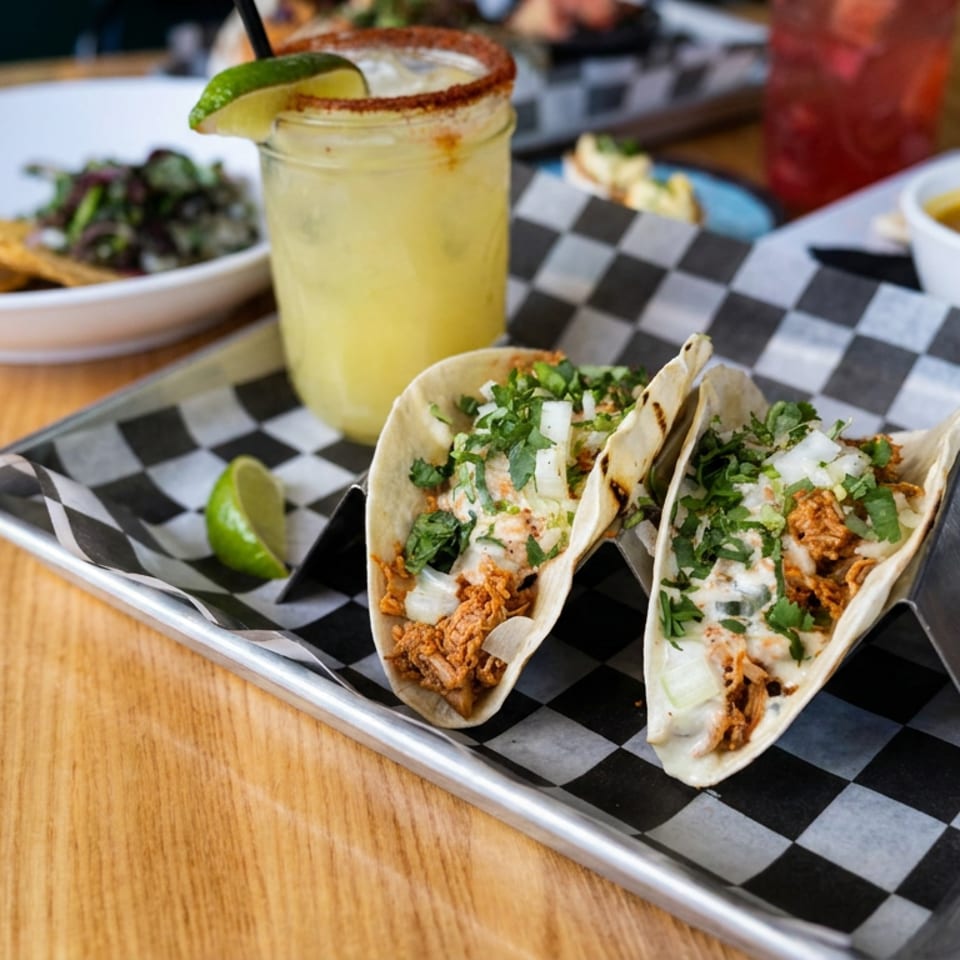 Two tacos topped with cilantro, onion, and crema sit on a checkered tray beside lime, salsa, chips, and a citrus drink.