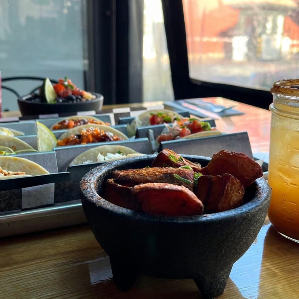 Tacos, roasted potatoes, and colorful drinks arranged on a restaurant table beside a sunlit window.