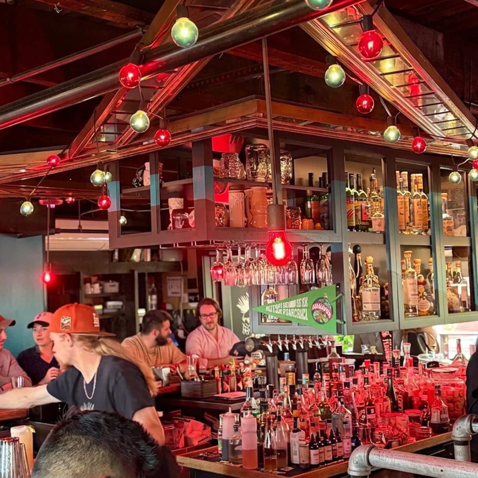 Lively restaurant bar with patrons, colorful string lights, liquor bottles, hanging glassware, and a glowing Legal Mezcal neon sign.
