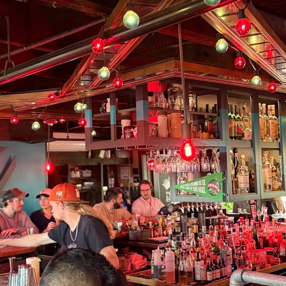 Bustling mezcal bar with colorful string lights, liquor shelves, bartenders, and patrons gathered around the counter.