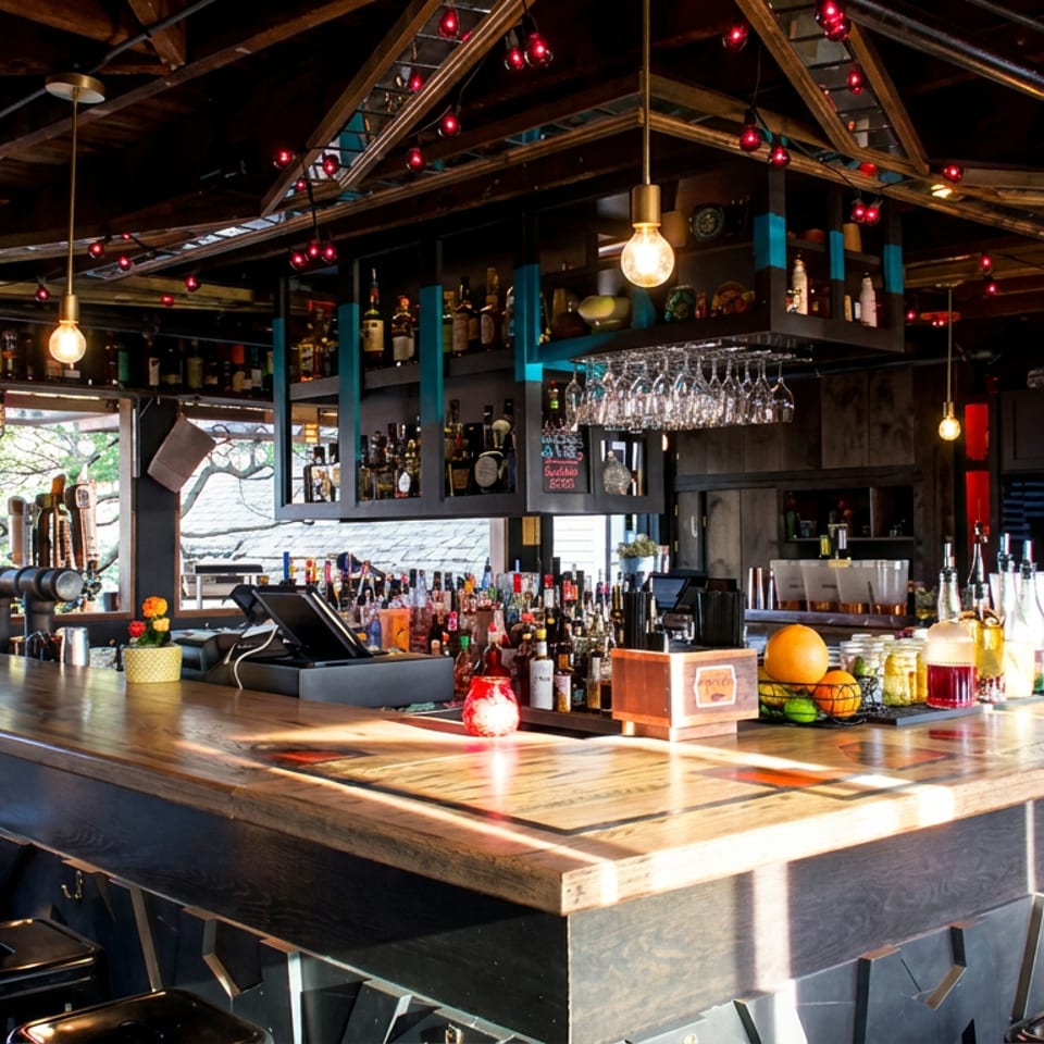 Rustic restaurant bar with liquor shelves, hanging glassware, pendant lights, bar stools, and sunlight through large windows.