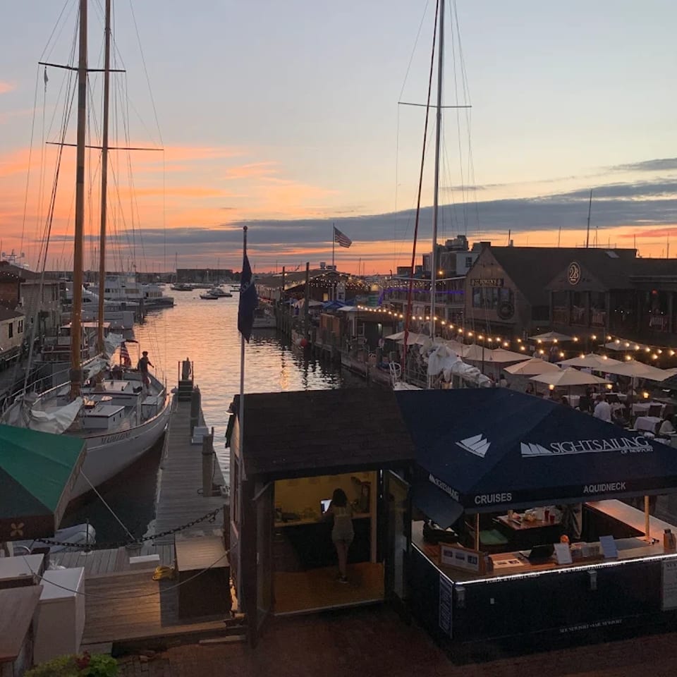 Sunset marina with sailboats, waterfront restaurants, outdoor umbrellas, string lights, and a sightseeing cruise kiosk along the dock.