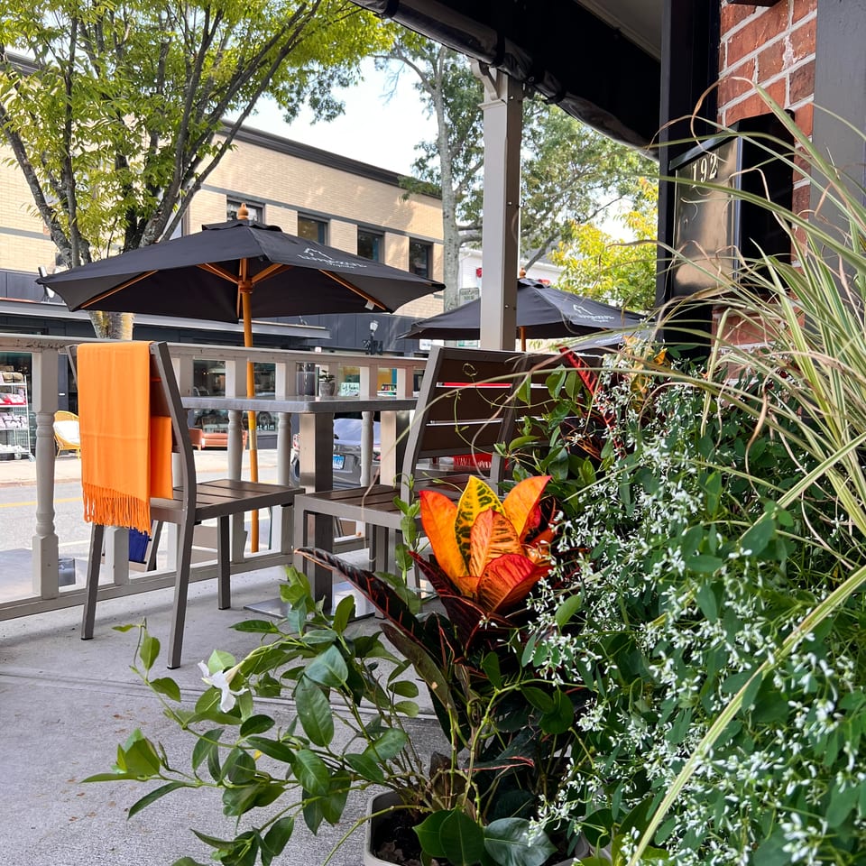 Restaurant patio with black umbrellas, orange-accented chairs, lush potted plants, and a brick facade.
