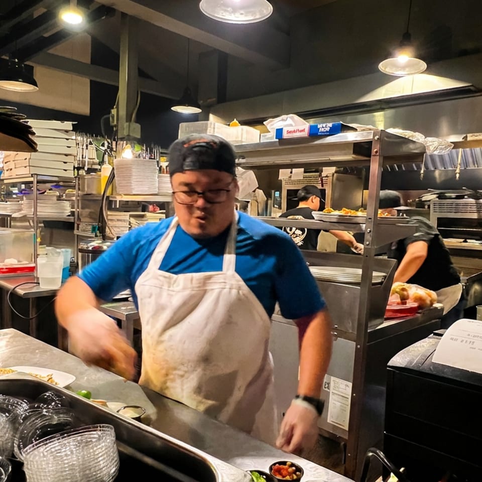 Restaurant kitchen worker in a blue shirt and apron prepares food at a busy stainless-steel counter.