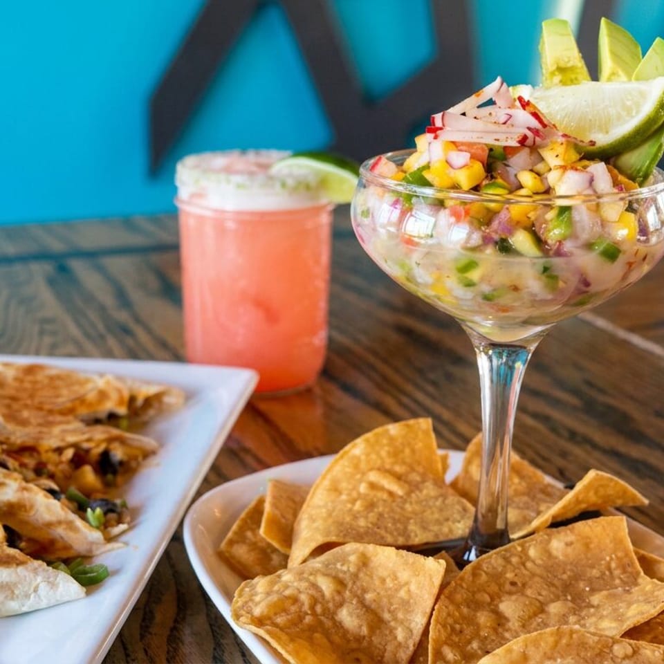 Mexican-style ceviche topped with lime and avocado, tortilla chips, quesadillas, and a pink drink on a wooden table.