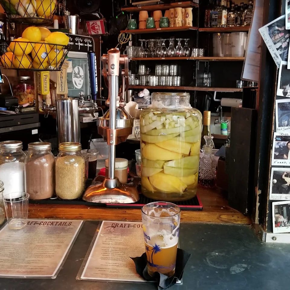 Busy craft cocktail bar with citrus basket, pickled fruit jar, drink, glassware, and handwritten photo display.
