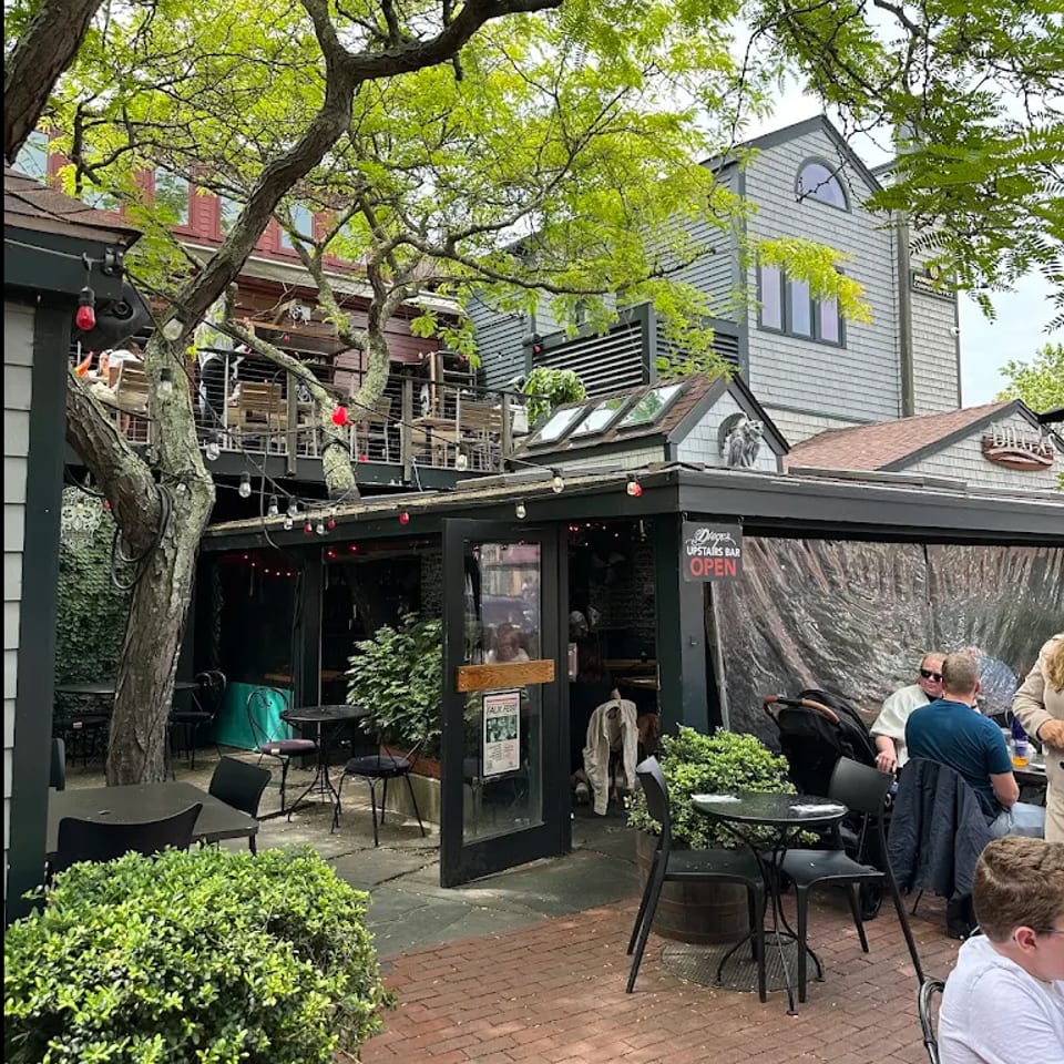 Tree-shaded restaurant patio with outdoor tables, string lights, people dining, and an entrance sign for the upstairs bar.