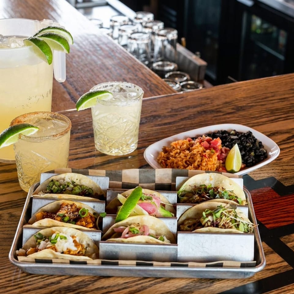 Tray of assorted tacos with lime wedges, rice and beans, margaritas, and a pitcher on a wooden restaurant bar.