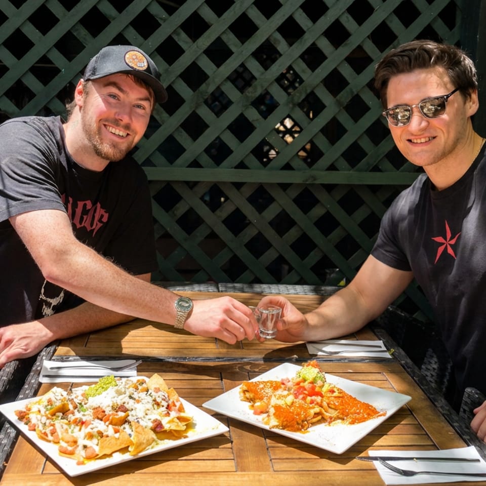 Two men toast with small glasses at a restaurant table beside plates of loaded nachos and saucy Mexican food.