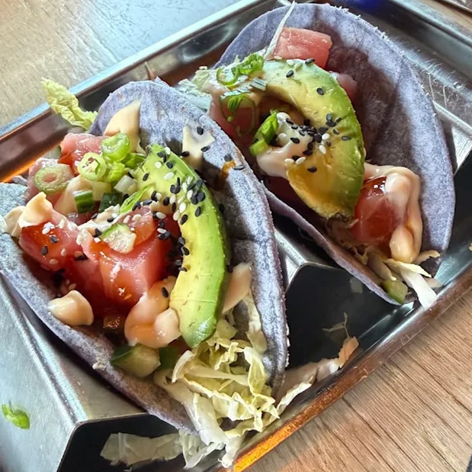Two soft blue-corn tacos topped with salmon, avocado, lettuce, green onions, sesame seeds, and creamy sauce on a metal tray.