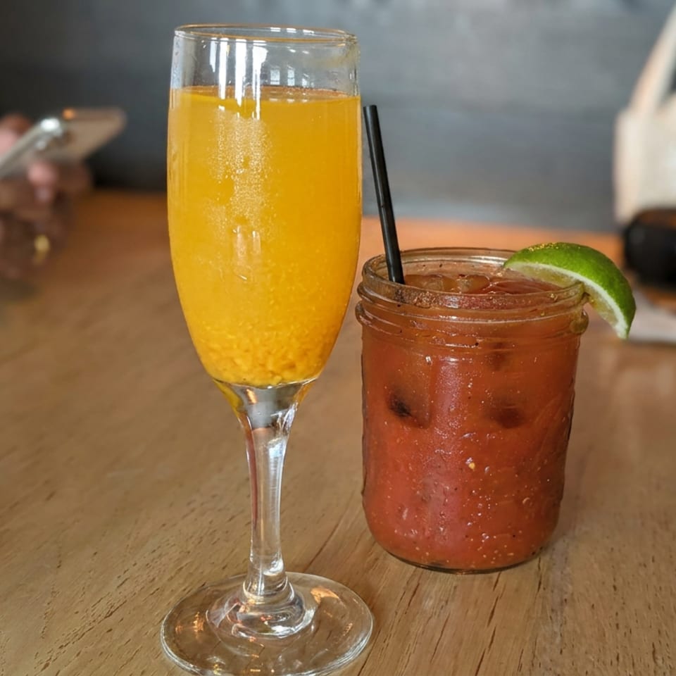 Orange sparkling drink in a flute and tomato-based cocktail with lime wedge on a restaurant table.